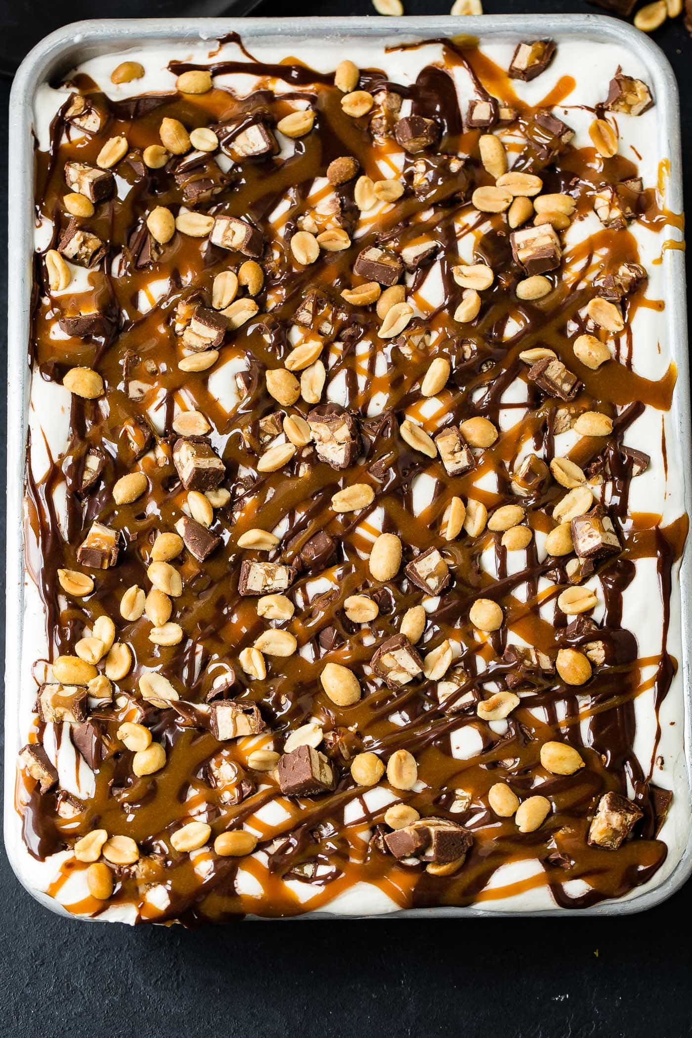 A pan of snickers ice cream cake. The top is covered with whipped cream, hot fudge, drizzles of caramel sauce, chopped snickers bars and salted peanuts