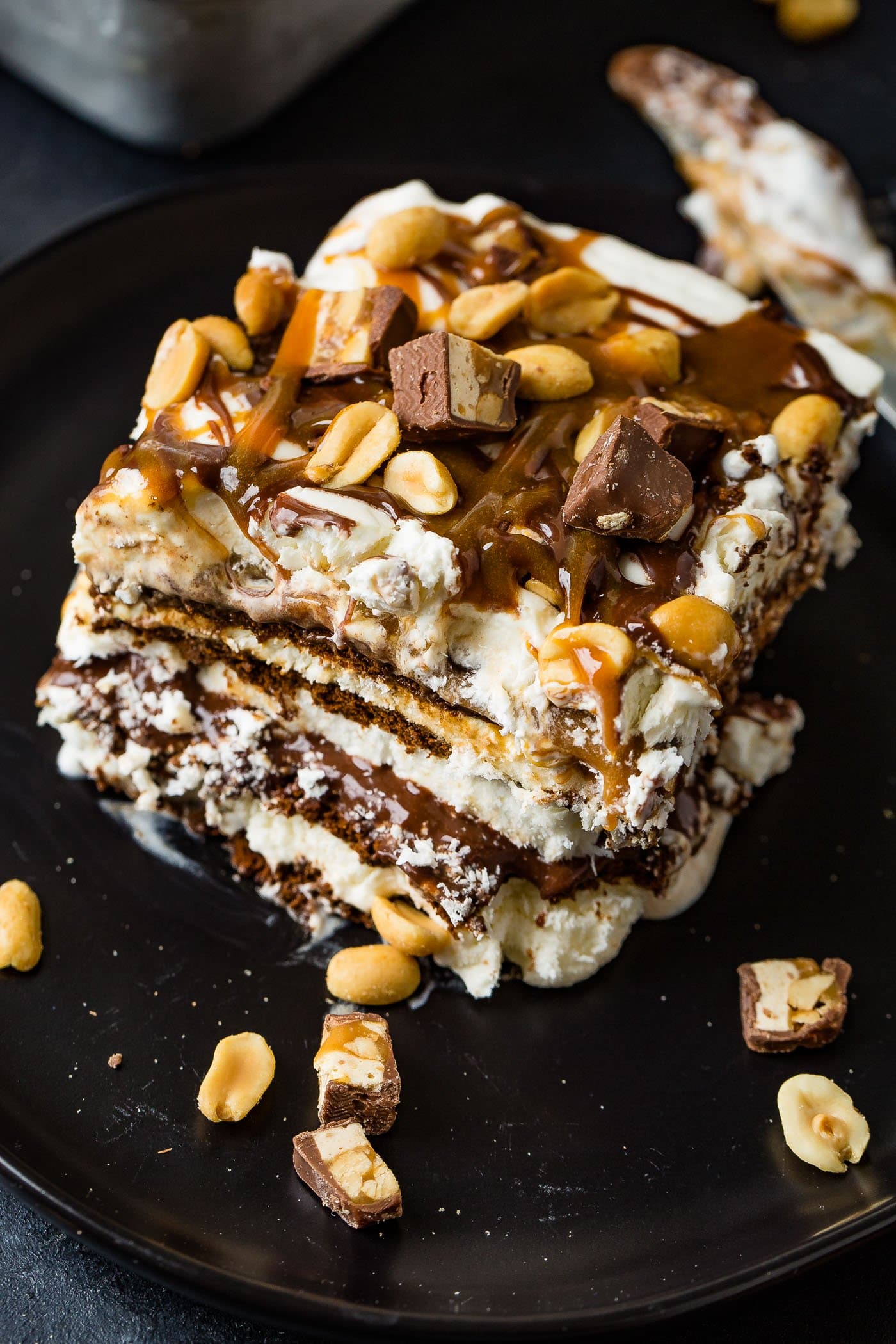 A piece of snickers ice cream cake on a dessert plate. The cake has layers of chocolate cake, ice cream, whipped cream, hot fudge, and caramel sauce. The top is covered with whipped cream, drizzles of hot fudge and caramel sauce, and salted peanuts. Chunks of snickers bars and peanuts are scattered around it.