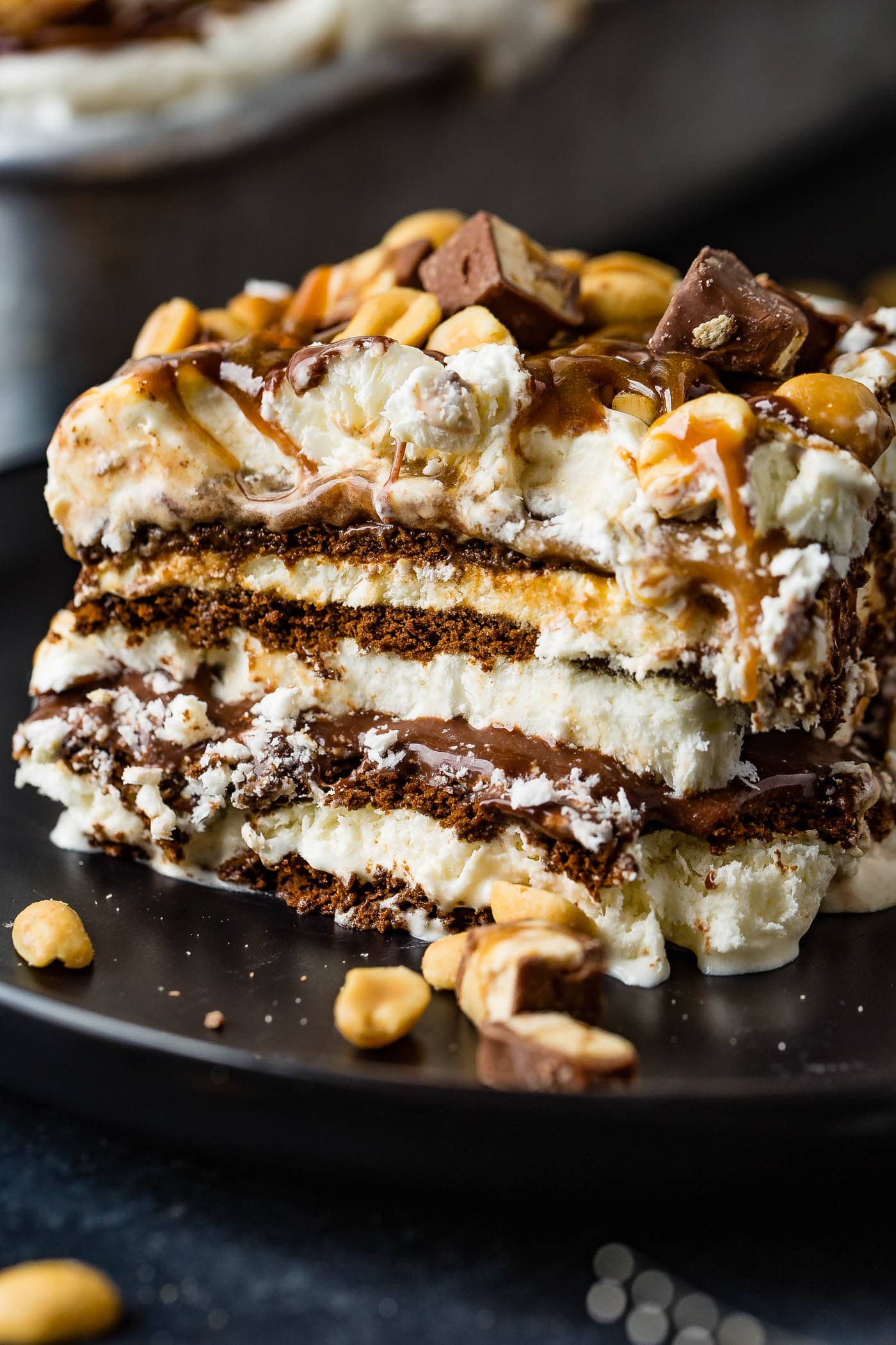 A square piece of snickers ice cream cake. There are layers of cake, ice cream, hot fudge, caramel sauce, snickers candy bars and whipped cream. The cake is topped with whipped cream, drizzles of hot fudge and caramel sauce and salted peanuts.